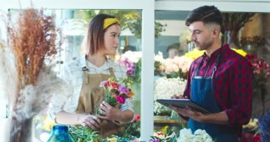 Çiçekçide yakışıklı bir adamla çalışan çekici beyaz kadın çiçekçi. Genç işçi tablet üzerinde yazıyor, envanter çıkarırken güzel kadın çiçek tutuyor. Ticaret konsepti.