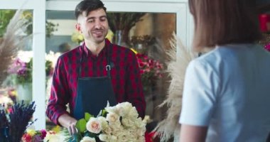 Çiçekçide çalışan önlük giyen çekici beyaz adam Florisr. Bayan müşteri çiçek buketini alıp dükkandan ayrılmaya geliyor. Ticaret, iş, girişimcilik, meslek..