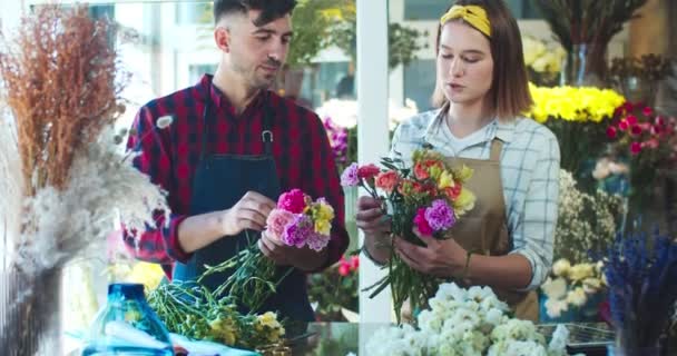 Jonge mannelijke en vrouwelijke bloemisten werken samen in een moderne bloemenwinkel. Vrolijke blanke man en vrouw die boeket van bloesems maken, met elkaar praten en lachen. Bedrijfsconcept. — Stockvideo