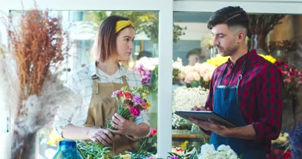 Florista feminina caucasiana atraente trabalhando com homem bonito na loja de flores. Jovem trabalhador está digitando em tablet, fazendo inventário, enquanto a mulher bonita está segurando flores. Conceito de comércio comercial. — Vídeo de Stock