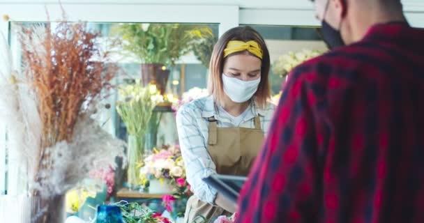 Молодые флористы и девушки в медицинских масках работают в цветочном магазине. Привлекательный мужчина и женщина разговаривают друг с другом, делают инвентаризацию и печатают на планшете. Технологии, концепция бизнеса. — стоковое видео