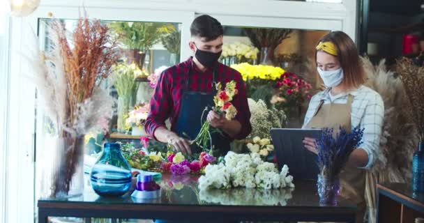 Aantrekkelijke blanke vrouwelijke bloemist werken met een knappe man in de bloemenwinkel. Jonge werkneemster typt op tablet, inventariseert terwijl knappe man boeket maakt. Handelsconcept. — Stockvideo