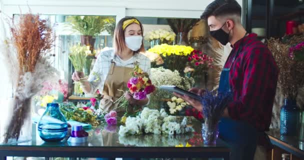 Hardwerkende mannelijke en vrouwelijke bloemisten werken in de bloemenwinkel. Knappe man is met behulp van tablet om de inventaris te nemen, terwijl mooie vrouw met bloemen en op zoek naar gadget, Business concept. Covid-19. — Stockvideo