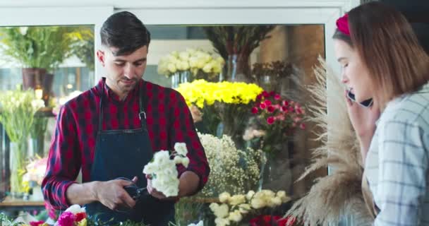 Handsome young male florist is cutting stems of flowers while his collegue calling client. Pretty Caucasian woman worker talking to customer and typing on tablet. Business, technology concept.