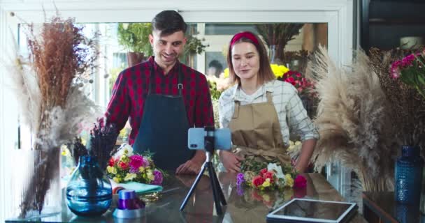 Jonge man en vrouw schieten video blog in de bloemenwinkel. Goed uitziende blanke bloemisten maken live stream conferentie op smartphone camera, het tonen van boeket aan kijkers, volgers. Sociale media. — Stockvideo