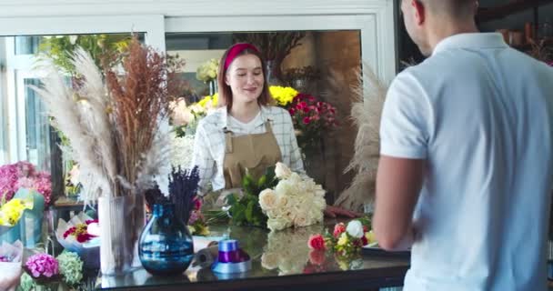 Piękna kobieta kwiaciarnia wita męskiego klienta i daje mu porządek w kwiaciarni. Przystojniak przychodzi do sklepu, żeby wziąć bukiet kwiatów, dać pieniądze i odejść. Biznes, koncepcja handlu. — Wideo stockowe