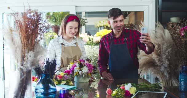 Young male and female florists calling to customer to discuss order. Pretty woman is showing bouquet while handsome man holding phone. Video face chat. Social media, technology, service concept. — Stock Video