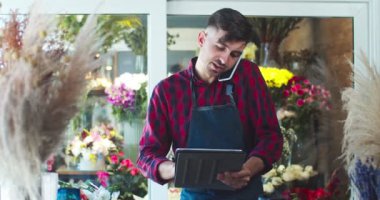 Çiçekçide çalışan önlüklü çekici bir gencin portresi. Yakışıklı beyaz erkek çiçekçi müşteriyi arıyor, müşteriyle konuşuyor ve tablet üzerinde yazıyor. İş, teknoloji, aletler konsepti.