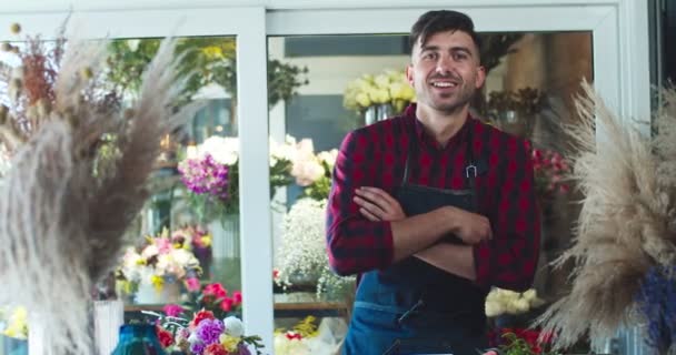 Jóképű fiatalember kötényben, egy modern virágboltban dolgozik. Jóképű fehér férfi virágárus keresztezi a karját, a kamerába néz és mosolyog. Üzleti, szakmai, vállalkozói koncepció. — Stock videók