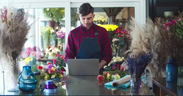 Modern çiçekçide dizüstü bilgisayar kullanan yakışıklı beyaz bir adam çalışıyor. Yakışıklı genç erkek çiçekçi çiçek envanterini tutuyor ve daktilo yazıyor. Ticaret, iş, girişimcilik kavramı. — Stok video