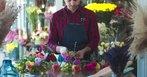 Close up de atraente jovem florista masculino vestindo avental, trabalhando na loja de flores. Bonito homem branco trabalhador fazendo buquê de flores. Negócio, conceito de empreendedorismo. — Vídeo de Stock