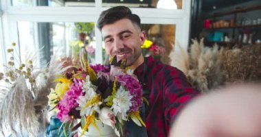 Yakışıklı beyaz adam çiçekçide dijital kamerayla video çekiyor. Elinde buket tutan yakışıklı erkek çiçekçi izleyicilere ve takipçilere çiçek açıyor. İş, günlük tutma kavramı.