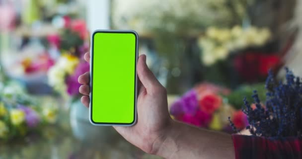 Primer plano del teléfono inteligente de mano masculina con croma key en la tienda de flores. Hombre mostrando teléfono celular con solapa verde. Flor borrosa fondo de la planta. Negocio, ocupación. — Vídeos de Stock