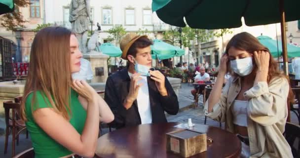 Feliz Caucásico amigos masculinos y femeninos posponer máscaras médicas y dar cinco altos al aire libre. Chico guapo y hermosas mujeres jóvenes sentadas en la terraza de la cafetería de buen humor. Concepto de cuarentena — Vídeo de stock