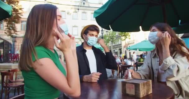 Jovenes amigos alegres sentados a la mesa en la terraza de la cafetería y quitándose las máscaras. Niño guapo caucásico en sombrero y hermosas hembras desinfectando las manos con spray al aire libre en la ciudad. Concepto de cuarentena — Vídeo de stock