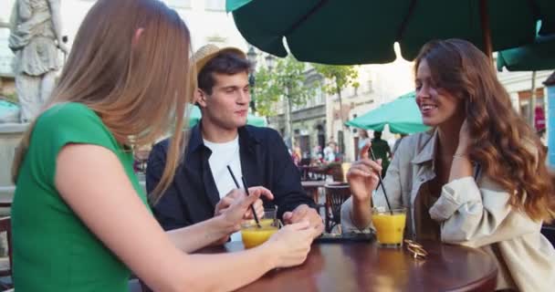 Кавказские счастливые друзья сидят на террасе кафетерия с напитками и общаются на открытом воздухе. Портрет радостного мальчика и красивых девушек, говорящих во время питья сока в городе. Концепция дружбы — стоковое видео