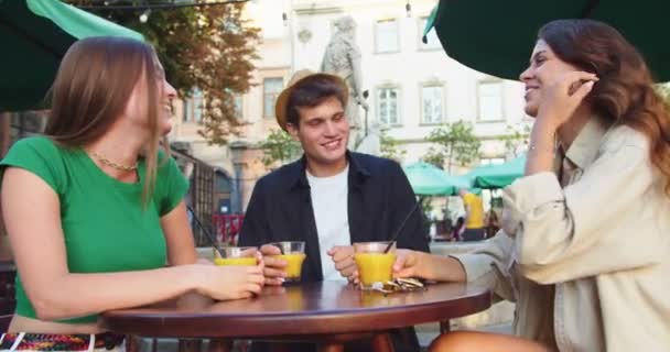 Retrato de alegres jóvenes amigos caucásicos sentados a la mesa afuera y bebiendo. Hombre guapo y mujeres hermosas sonriendo y levantando vasos con bebidas en la terraza de la cafetería en la ciudad. Concepto de ocio — Vídeo de stock