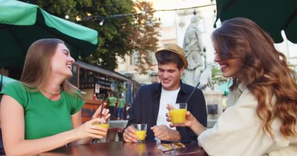 Fröhliche kaukasische junge Leute sitzen am Tisch auf der Restaurantterrasse und nippen an Getränken. Schöner Mann und schöne Frauen lachen und heben Gläser mit Getränken im Freien in der Stadt. Freizeitkonzept — Stockvideo