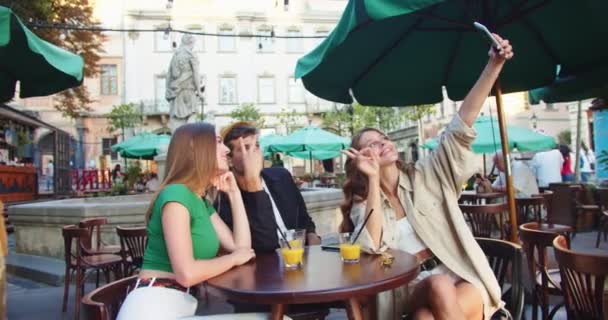 Щасливий хлопець з Кавказу і вродливі дівчата роблять фотографії на смартфоні, сидячи за столиком на подвір'ї. Радісна молодь фотографує мобільний телефон у місті. Дивна концепція — стокове відео