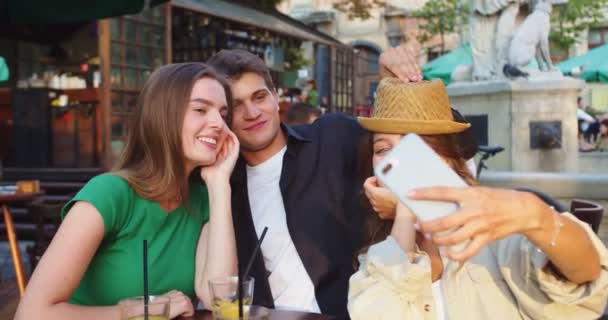 Ritratto di ragazzo bello caucasico gioioso e belle ragazze che fanno foto selfie su smartphone mentre si siedono a tavola all'aperto. Amici felici che scattano foto al cellulare in mensa. Concetto di tempo libero — Video Stock