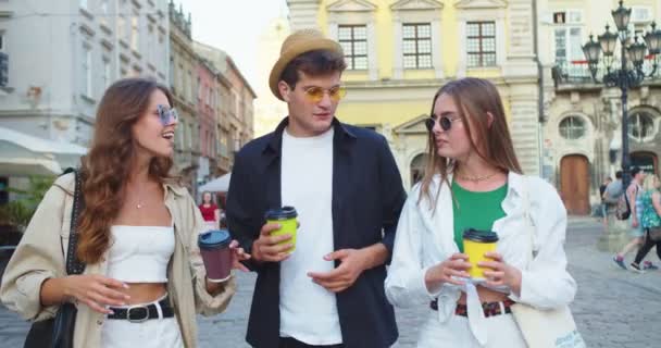 Portré boldog kaukázusi barátokról, akik kávét isznak, miközben a városban sétálnak és beszélgetnek. Jóképű fiú sapkában és gyönyörű lányok napszemüvegben beszélgetnek és mosolyognak az utcán. Szabadidős koncepció — Stock videók