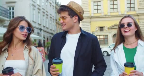 Portré vidám kaukázusi barátokról, akik elvitelre kávéznak, miközben a városban sétálnak és beszélgetnek. Jóképű férfi kalapban és gyönyörű lányok napszemüvegben beszélgetnek és mosolyognak az utcán.. — Stock videók
