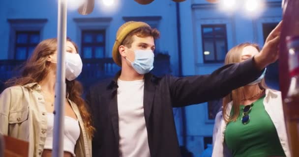 Retrato de amigos felices comprando café para llevar en la calle por la noche. Hombre caucásico guapo en sombrero y chicas en máscaras médicas tomando café en la ciudad. Gente levantando copas al aire libre. Concepto Amigos — Vídeos de Stock