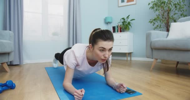 Plan lateral medio de fuerte hermosa chica de fitness en ropa deportiva atlética está haciendo ejercicios de tablón mientras usa un cronómetro en su teléfono. La hembra está entrenando en casa. Gente y concepto de estilo de vida activo. — Vídeo de stock