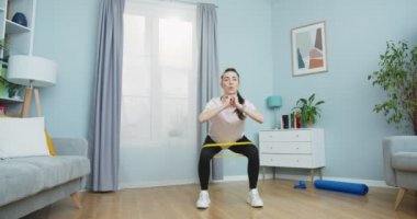 Yetişkin bir kadının evde oturma odasında direnç bandıyla oturması, sağlıklı bir yaşam tarzı için mekik çekmesi, kalçaları için spor yapması. Spor ve zindelik.