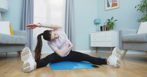 Plano medio de la hermosa mujer confiada en pose dividida que se extiende sobre una alfombra azul en el piso de la sala de estar utilizando un teléfono inteligente con entrenamiento en línea. Deporte y fitness. Concepto de entrenamiento, entrenamiento y bienestar. — Vídeos de Stock