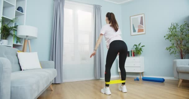 Back view of young attractive woman exercising with resistance power band, doing fitness workout for healthy lifestyle. Girl squats indoor in living room, doing sit ups exercises. — Stock Video