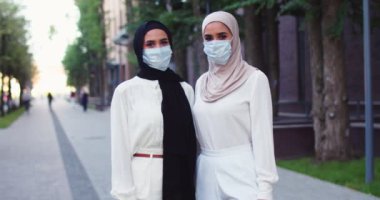 Geleneksel türbanlı ve tıbbi maskeli iki güzel, mutlu Arap kadının kameraya baktığı bir plan. Müslüman, neşeli, tesettürlü kadınlar. Şehirde İslamcı kadınlar.