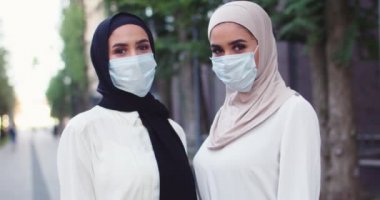 Geleneksel türbanlı ve tıbbi maskeli, kameraya bakan iki güzel mutlu Arap kadının portresi. Müslüman, neşeli, tesettürlü kadınlar. Şehirde İslamcı kadınlar.