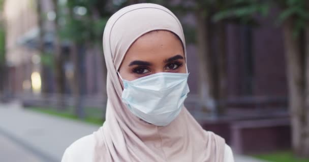 Primer plano retrato de la joven musulmana hermosa mujer en el pañuelo de cabeza tradicional y máscara médica mirando a la cámara. Primer plano de la mujer árabe con hermosos ojos en hijab en la calle. — Vídeos de Stock