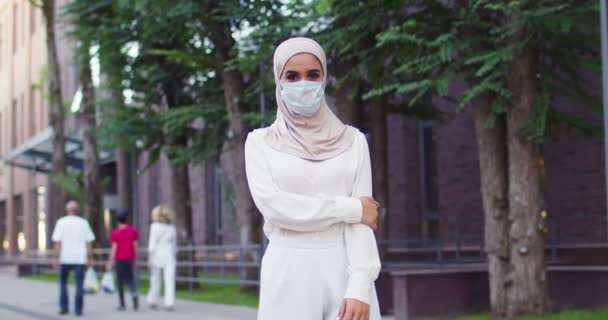 Plano medio de la joven musulmana hermosa mujer en el pañuelo de cabeza tradicional y máscara médica mirando a la cámara con ropa blanca con clase. Primer plano de la mujer árabe con hermosos ojos en hijab en la calle. — Vídeo de stock