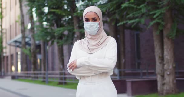 Środkowy plan młodej muzułmańskiej pięknej kobiety w tradycyjnej chustce i masce medycznej przechodzącej przez ramiona i patrzącej w kamerę. Zbliżenie arabskiej kobiety z pięknymi oczami w hidżabie na ulicy. — Wideo stockowe