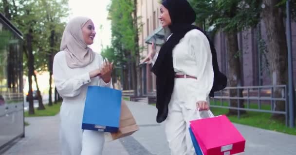 Plano medio de tiro de dos jóvenes árabes hermosas mujeres en hiyabs. Mujer musulmana sonriendo, discutiendo, hablando, caminando en la calle de una ciudad, sosteniendo bolsas de compras en las manos y saltando de felicidad. — Vídeo de stock