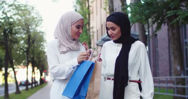 Foto de plan medio de dos mujeres jóvenes árabes hermosas en traje blanco casual y hiyabs blancos y negros. Sonriendo, discutiendo, hablando, caminando en la calle de una ciudad, sosteniendo bolsas de compras en las manos. — Vídeos de Stock