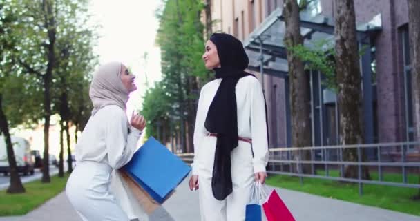 Plano medio de dos jóvenes mujeres árabes en pañuelos tradicionales sonriendo, discutiendo, hablando, caminando por la calle, sosteniendo bolsas de compras en las manos y saltando de felicidad después de compras exitosas. — Vídeo de stock