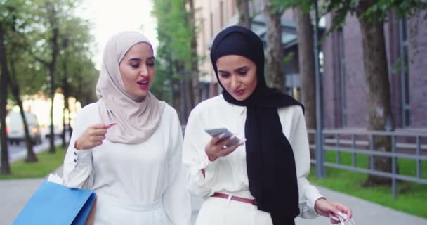 Środkowy plan dwóch arabskich kobiet w tradycyjnych chusteczkach do głowy dokonujących zakupów online, dokonujących zakupów za pomocą smartfona. Piękne arabki w hidżabach w mieście. Islamskie szczęśliwe panie w eleganckim białym stroju. — Wideo stockowe