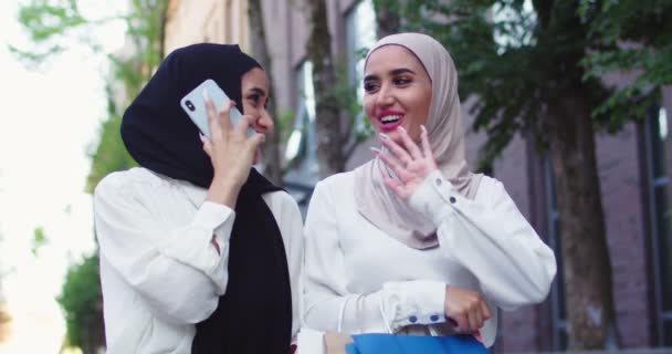 Close up portraits of two Arab smiling women in traditional headscarf strolling at city, talking on phone and carrying bags from shop. Female hijab speaking on cellphone after shopping and laughing. — Stock Video