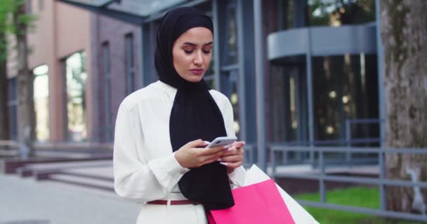 Arabska młoda kobieta w tradycyjnych chusteczkach patrzy na telefon idący ulicą. Ładna muzułmanka w hidżabach czytająca wiadomości, przewijająca złe wieści na smartfonie po zakupach. — Wideo stockowe