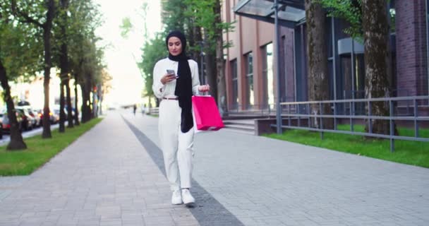 Арабська вродлива жінка в традиційних головних уборах дивиться на телефонну прогулянку по вулиці. Досить муслімна жінка в бесідах, надсилає повідомлення, дзвонить, говорить по смартфону після покупки.. — стокове відео