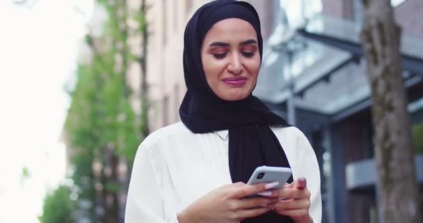 Arabic woman in traditional headscarves looking at phone walking at stree and smiling. Pretty muslim female in hijabs reading messages, texting, browsing Internet on smartphone after shopping. — Stock Video