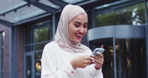 Arab nő hagyományos fejkendőben nézi a telefont, ahogy Stree-n sétál és boldogan nevet. Csinos muszlim nő hidzsáb olvasás üzenetek, SMS-ezés, böngészés az interneten okostelefonon vásárlás után. — Stock videók