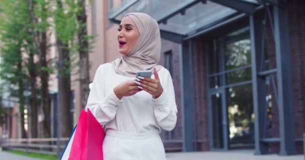 Arabska kobieta w tradycyjnych chustkach patrzy na telefon spacerując po Stree i szczęśliwy śmiejąc się. Pretty muzułmanka w hijabs czytanie wiadomości, SMS-y, przeglądanie Internetu na smartfonie po zakupach. — Wideo stockowe