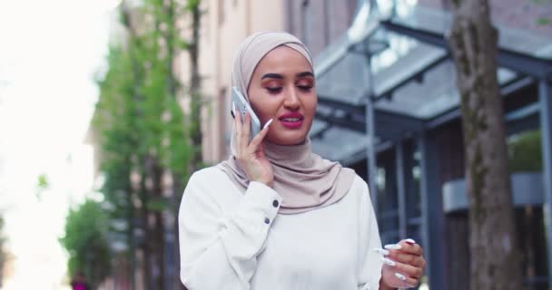 Primer plano de mujer árabe hermosa en pañuelos tradicionales hablando por teléfono caminando en la calle. Bastante musulmana hembra en hijabs haciendo llamada en el centro de la ciudad después de ir de compras. — Vídeos de Stock
