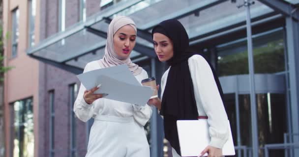 Two young confident Arabic beautiful women in traditional headscarves passing documents outdoors on sunny day and talking. Professional coworkers discussing cooperation. Teamwork, business, lifestyle. — Stock Video