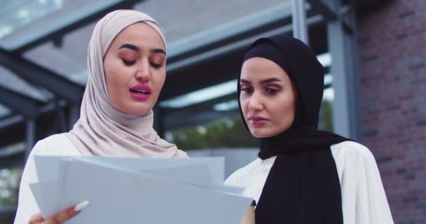 Primer plano de dos mujeres musulmanas de negocios en pañuelos tradicionales discutiendo documentos al aire libre, hablando, bebiendo café. Colaboradores profesionales discutiendo cooperación. Trabajo en equipo, negocios, estilo de vida. — Vídeo de stock