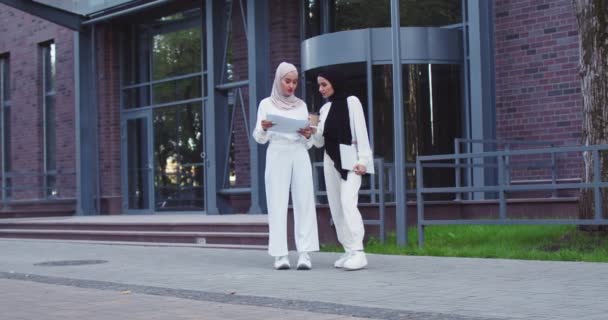 Δύο αισιόδοξες μουσουλμάνες όμορφες γυναίκες με παραδοσιακές μαντίλες συζητούν έγγραφα σε εξωτερικούς χώρους, μιλάνε, πίνουν καφέ. Επαγγελματίες συνάδελφοι συζητούν για συνεργασία. Ομαδική εργασία, επιχείρηση, τρόπος ζωής. — Αρχείο Βίντεο